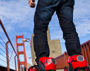 A person wearing red, motorized roller shoes stands on a sidewalk near the Golden Gate Bridge, with the iconic bridge structure visible in the background.