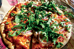 A close-up of a thin-crust pizza topped with tomato sauce, melted cheese, red onion slices, and a generous amount of fresh arugula. The pizza is cut into slices and served on brown parchment paper.