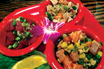 Three red bowls filled with colorful poke salads, including diced fish, vegetables, and herbs, are arranged on a red tray with a purple flower garnish and lemon slice nearby.
