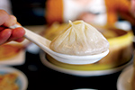 A close-up of a steamed dumpling in a white spoon, held in the foreground with a blurred person and plate in the background.