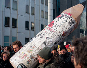 A man carries a large pencil prop covered in newspaper clippings through a crowd of protesters on a city street, with tall buildings in the background.