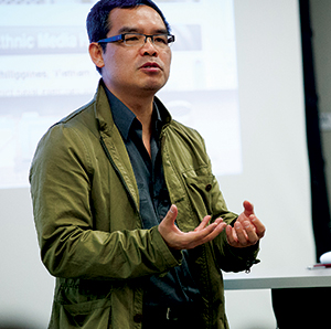 A man wearing glasses and an olive-green jacket gestures with his hands while speaking in front of a screen during a presentation.