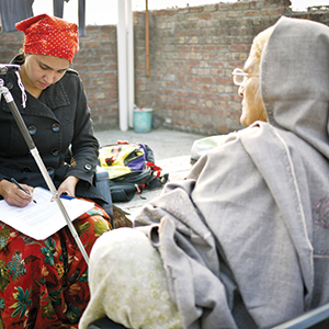 A woman in a red headscarf interviews an elderly woman wrapped in a gray shawl, writing notes on a clipboard outdoors near a brick wall.