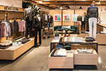 A modern clothing store displays neatly arranged mens apparel on racks and tables, with jackets, shirts, and pants, set in a well-lit space with a seating area and framed art on the back wall.