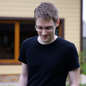 A man with short brown hair and glasses, wearing a black t-shirt, looks down and smiles slightly while standing outside in front of a building with yellow siding and a window.