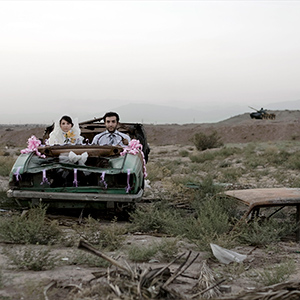 A bride and groom sit in a decorated, roofless car surrounded by dry, grassy terrain. There is an abandoned car and a helicopter in the background. The scene appears desolate and unconventional for a wedding.