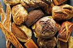 A variety of artisanal breads, including baguettes, round loaves, and rolls, arranged closely together on a dark surface.