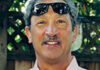 A man with gray hair and a mustache smiles outdoors, wearing sunglasses on his head and a light-colored shirt. A wooden fence and greenery are in the background.