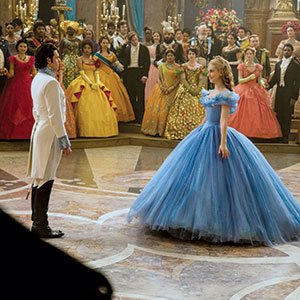 A woman in a blue ball gown and a man in a white suit stand facing each other in a grand ballroom, surrounded by elegantly dressed guests and ornate decorations.