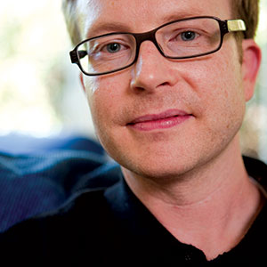 A close-up of a man with short light brown hair and glasses, wearing a dark shirt. He is looking at the camera with a neutral expression, with a soft-focus background.