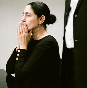 A woman with dark hair tied back sits with her hands clasped near her face, appearing thoughtful or concerned. She wears a black long-sleeve top with gold buttons. Part of a man in a suit is visible beside her.