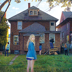 A blonde woman in a blue jacket stands in a grassy backyard facing the camera, while three people and a child stand near the back of a brown brick house under a clear sky.