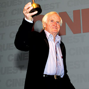 An older man with white hair holds up a trophy while standing in front of a gray background with the word CINEQUEST repeatedly printed on it. He is wearing a dark jacket over a light shirt.