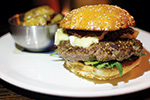 A close-up of a sesame seed burger bun filled with a large beef patty, lettuce, cheese, and sauce, served on a white plate with a side of pickles in a small metal bowl.