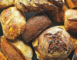 An assortment of rustic breads including round, oval, and rectangular loaves with crusty golden-brown exteriors, arranged closely together.
