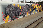 A mural along a train track features a large, black, abstract creature entwined with red lines and surrounded by yellow, coin-like shapes on a grey background.