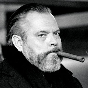 A bearded man with slicked-back hair stares intently at the camera while holding a large cigar in his mouth. He is wearing a dark coat. The background is out of focus.