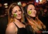Two young women with shamrock face paint smile at the camera in a lively, crowded bar. Both have long hair and are dressed casually. Neon signs and blurred people are visible in the background. metroactive logo is at the bottom right.