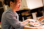A woman in a checkered shirt is smiling while pouring sauce from a saucepan onto a dish in a kitchen.
