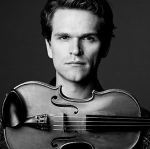 A man with short hair looks directly at the camera, holding a violin upright in front of his face. The image is in black and white, with a dark background.