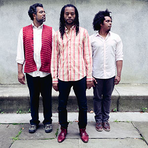 Three men stand in front of a concrete wall. The man in the center faces forward, while the men on each side face toward him. They are dressed casually in button-down shirts and slacks.