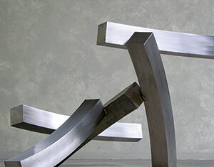 A modern abstract sculpture made of three large, curved, metallic beams intersecting and balancing on a plain concrete floor against a gray textured background.