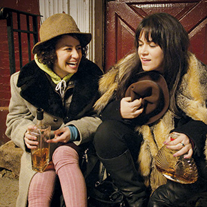 Two women sit on outdoor steps at night, wearing winter coats and hats, smiling and holding bottles of liquor, appearing to enjoy each others company. One woman is laughing while the other looks down.