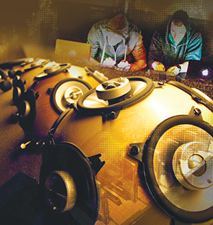 A row of large, round speakers in the foreground, with two people in hoodies working on laptops in a dim, atmospheric setting in the background. The image has a warm, golden hue and a tech-focused ambiance.