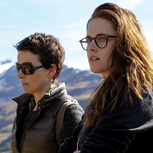 Two women wearing glasses stand outdoors in front of a scenic, mountainous background. One has short dark hair and sunglasses; the other has long brown hair and wears clear eyeglasses. Both look into the distance.