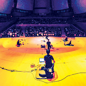 Several people sit in a wide circle on a brightly lit stage facing an audience in a large auditorium. Electronic equipment and cables are arranged around them on the floor.