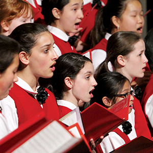 A group of girls wearing white shirts and red vests sing in a choir, holding red folders. Their mouths are open mid-song and they appear focused on their performance.