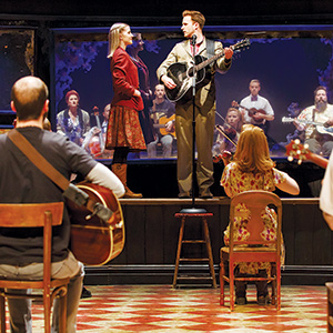 A man stands on a stool playing guitar and singing to a woman on stage, while other musicians sit and play guitars and string instruments, watching them perform. The scene is set in a theater with warm lighting.