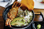 A bowl of rice topped with fried soft-shell crab, sliced avocado, shredded vegetables, a crunchy chip, and sprinkled with seaweed flakes, served with dipping sauce and wasabi on the side.
