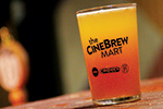 A pint glass with a gradient yellow to red drink sits on a wooden surface. The glass reads the CINEBREW MART along with smaller logos. The background is blurred.