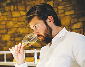A man with a beard and neatly styled hair, wearing a white shirt, sits indoors against a stone wall and smells a glass of white wine, appearing to concentrate on its aroma.