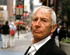 An older man with short gray hair and a serious expression stands outdoors in a city, wearing a white shirt and dark jacket. People and buildings are blurred in the background.