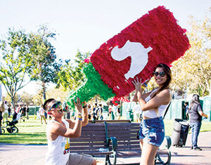 Two people outdoors hold a large, red piñata shaped like a sriracha bottle; one person kneels, pretending to drink from it, while the other stands and smiles, both wearing sunglasses and casual summer clothes.
