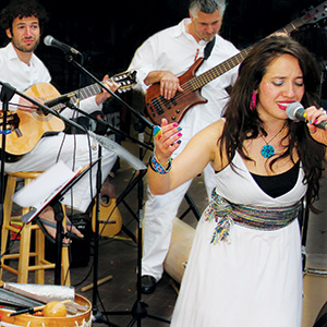 A woman sings passionately into a microphone while two men behind her play guitar and bass. All three musicians are dressed in white. Musical instruments and equipment are visible around them.