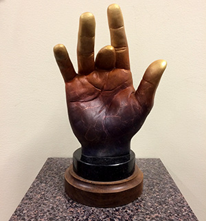 A sculpture of an upright hand with the thumb, index, and pinky fingers extended, while the middle and ring fingers are folded down. The hand is bronze-colored with gold fingertips, mounted on a round base.
