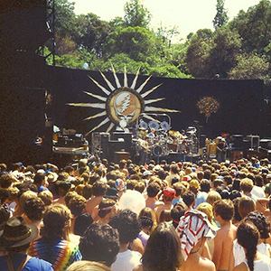 A large crowd watches a band perform on an outdoor stage with a black backdrop featuring a sunburst design and a skull with a lightning bolt. Trees and daylight are visible in the background.