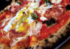 Close-up of a pizza topped with tomato sauce, melted cheese, slices of salami, fresh basil leaves, and a baked egg with a runny yolk on a charred, bubbly crust.