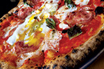 Close-up of a pizza topped with tomato sauce, melted cheese, slices of salami, fresh basil leaves, and a baked egg with a runny yolk on a charred, bubbly crust.