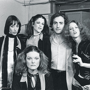 Five people in stylish 1970s clothing pose together indoors, with serious and expressive looks. Three women stand in back, one man and one woman are in front. The setting appears to be backstage with visible wires and equipment.
