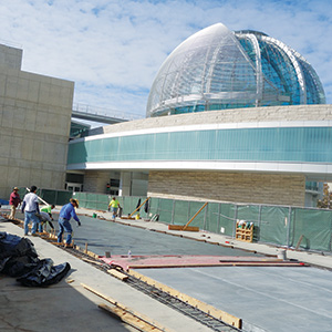Several construction workers pour and smooth concrete outside a modern building with a large glass dome and curved facade under a partly cloudy sky.