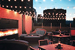Cozy outdoor patio at dusk with string lights overhead, a lit fireplace on the left, cushioned seating, and several tables and chairs arranged for dining or relaxation.