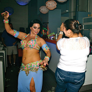 A belly dancer in a colorful costume performs enthusiastically in a dimly lit room while a woman in casual clothes stands nearby, appearing to enjoy the dance.