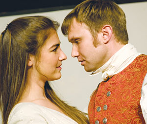 A woman and a man in period costumes stand close, gazing intently into each other’s eyes, suggesting a romantic or dramatic moment. The man wears a red patterned vest and white collar; the woman wears a light dress.