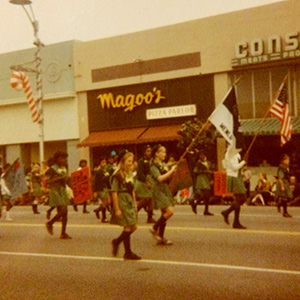 A group of girls in green uniforms marches in a parade, carrying flags and signs in front of Magoos Pizza Parlor on a city street. Spectators watch from the sidewalk.