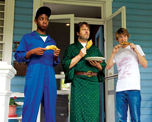 Three people stand on a porch eating pastries. The person on the left wears a blue jumpsuit and cap, the middle person wears a green robe and holds a plate, and the person on the right wears a white T-shirt and jeans.