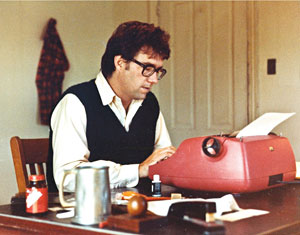 A man wearing glasses and a dark vest sits at a desk typing on a red typewriter. Various office supplies, a mug, and a bottle are on the wooden desk. A plaid shirt hangs on the wall behind him.